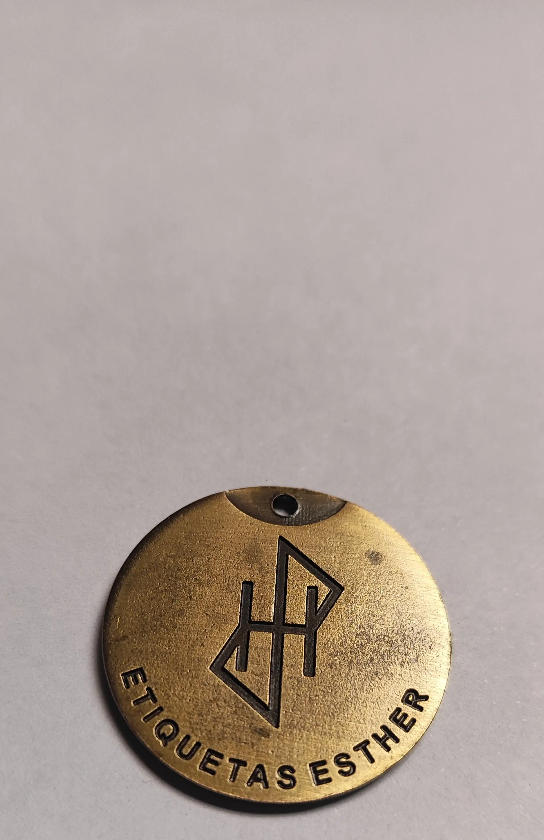 Medalla redonda de zamak en bronce tamboreado, muy ampliada, con el monograma y el nombre de la marca grabados en hueco y el grano del metal a la vista
