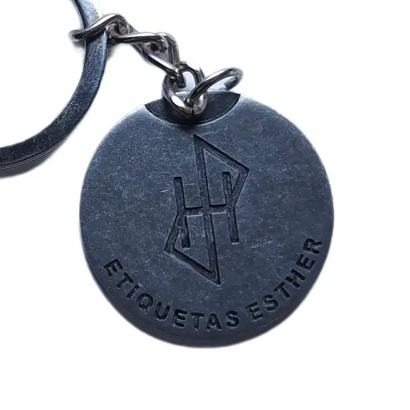 La misma medalla redonda de zamak en gris pistola, en primer plano, con el monograma grabado en hueco
