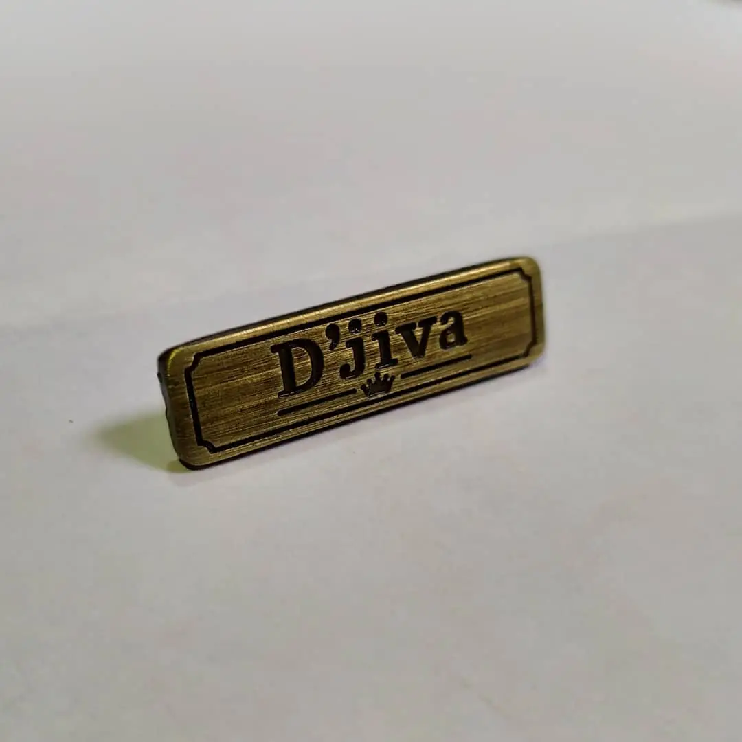 Etiqueta metálica en bronce tamboreado con marco en relieve y dos perforaciones para coser