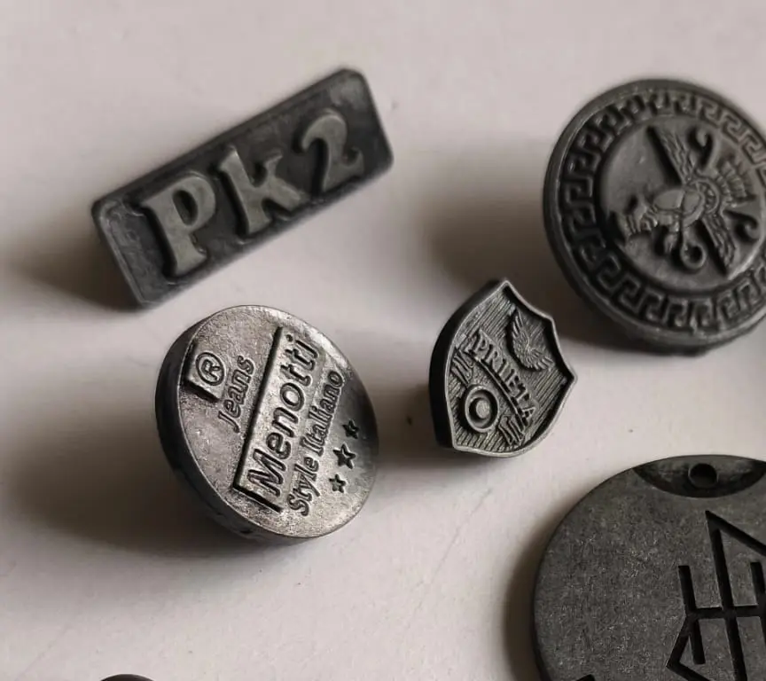 Cuatro insignias de zamak en gris pistola: una rectangular, dos redondas y un escudo