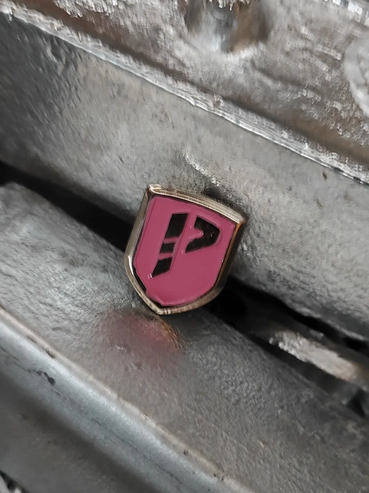 Pin de zamak en forma de escudo, con el logo de una marca sobre esmalte rosado, apoyado sobre lingotes de zamak