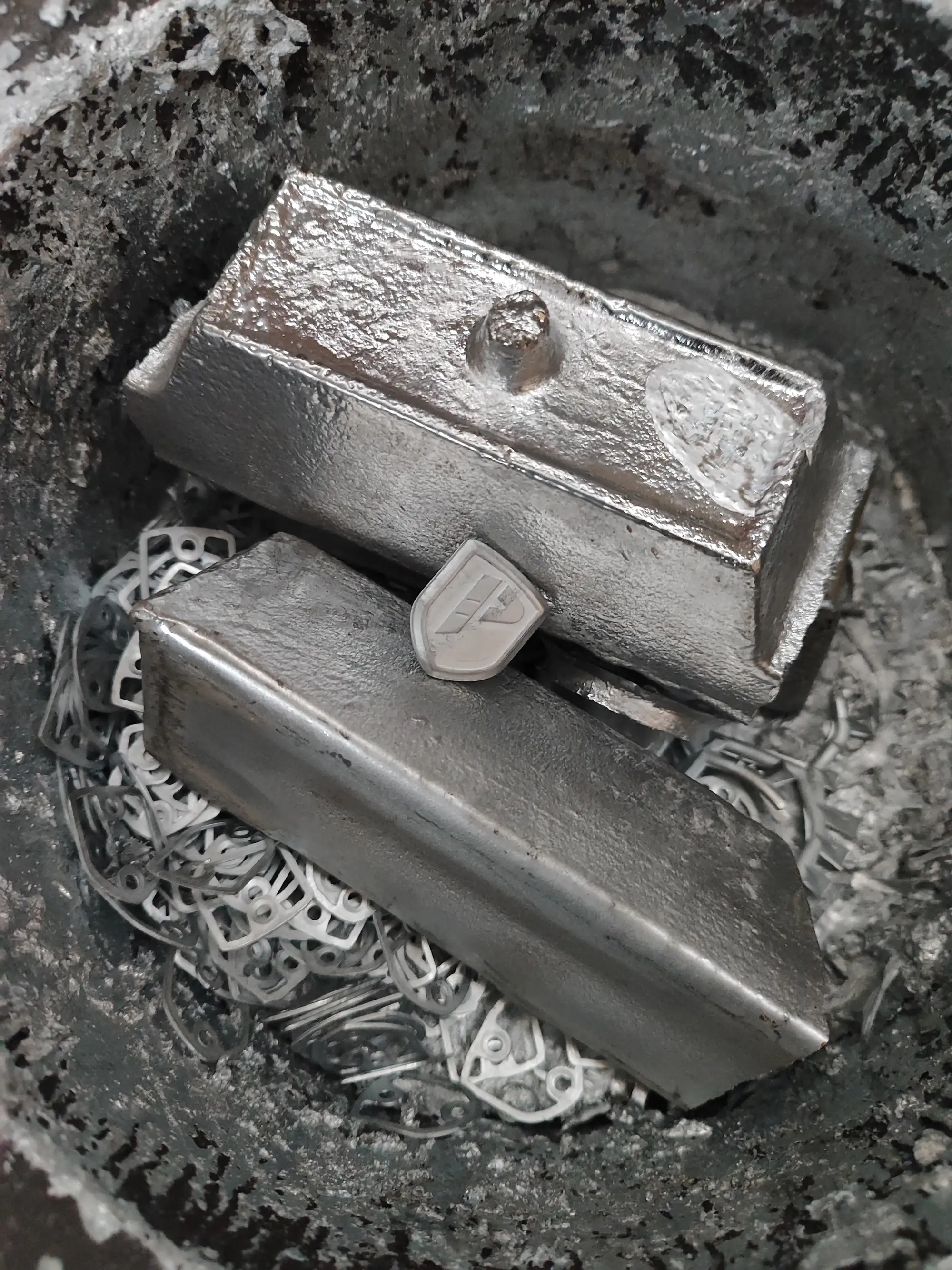Lingotes de zamak dentro del crisol, sobre recortes de metal que vuelven a fundirse, con una pieza recién fundida entre ellos