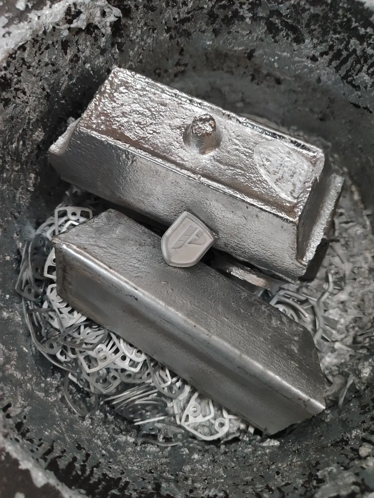 Lingotes de zamak dentro del crisol, sobre recortes de metal que vuelven a fundirse, con una pieza recién fundida entre ellos
